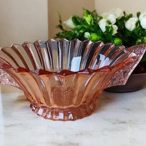 Peut inclure: Un bol en verre rose vintage avec un bord festonné et des poignées décoratives. Le bol est posé sur une surface en marbre blanc, avec un arrière-plan flou de fleurs blanches dans un bol marron.