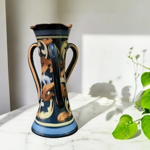 Puede incluir: Un jarrón decorativo de cerámica, alto, con dos asas. El jarrón presenta una base y un borde azul oscuro, con una franja azul más clara. El cuerpo está adornado con patrones arremolinados marrones, beige y crema. El jarrón mide unos 30 cm de alto.