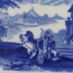 May include: A blue and white ceramic plate featuring a classical scene. A horse-drawn chariot with figures is depicted in the foreground, with a mountainous landscape and architectural elements in the background. The design is in a traditional blue and white color scheme.