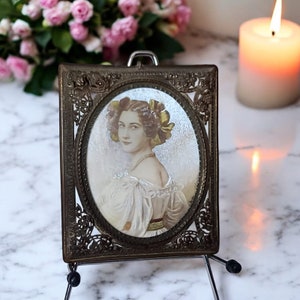 May include: Antique portrait in an ornate bronze frame. The oval portrait features a woman in a white dress with yellow accents. The frame has a decorative filigree design. The portrait is displayed on a small metal easel.