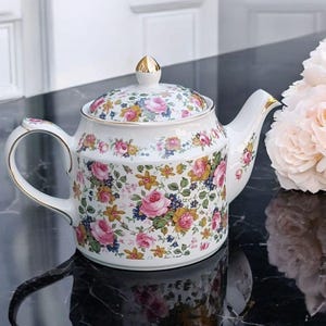 Può includere: Teiera in ceramica bianca con un motivo floreale di rose rosa, fiori gialli e dettagli blu. La teiera ha un manico, un beccuccio e un pomello dorati. Un bouquet di rose rosa chiaro è accanto.