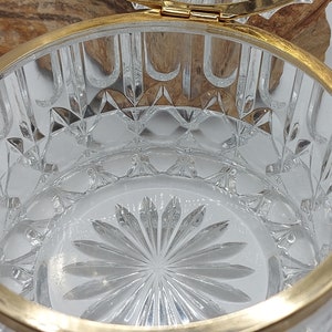 Puede incluir: Un recipiente de vidrio tallado transparente con un borde y bisagra de metal dorado. El vidrio presenta un patr&oacute;n de diamantes y un dise&ntilde;o floral en la base. La tapa est&aacute; abierta, revelando el interior. El recipiente es probablemente para joyas o objetos peque&ntilde;os.