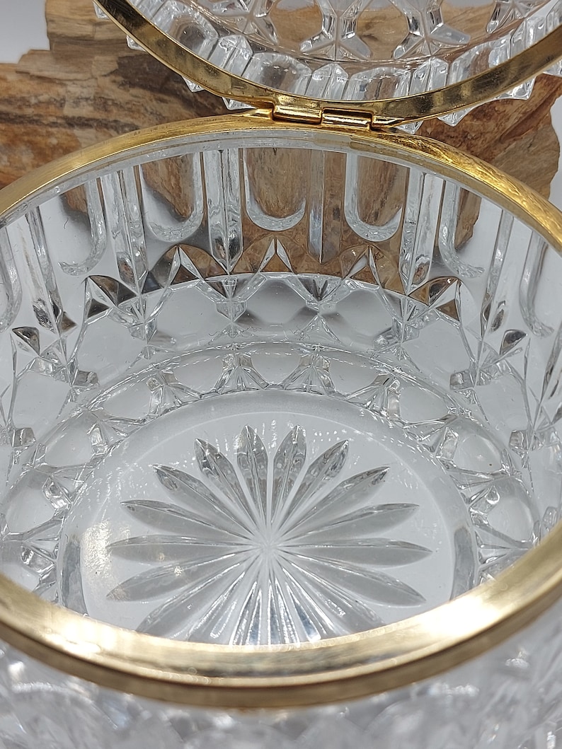 Puede incluir: Un recipiente de vidrio tallado transparente con un borde y bisagra de metal dorado. El vidrio presenta un patr&oacute;n de diamantes y un dise&ntilde;o floral en la base. La tapa est&aacute; abierta, revelando el interior. El recipiente es probablemente para joyas o objetos peque&ntilde;os.