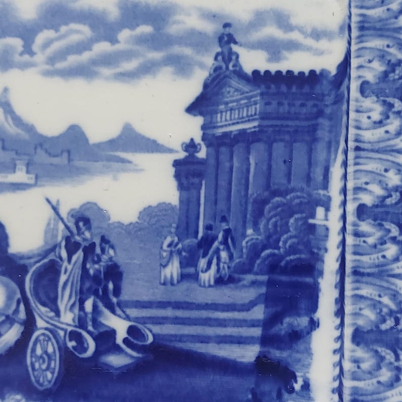 May include: A section of a blue and white ceramic plate featuring a detailed landscape scene. The design includes figures, architectural elements, and a chariot, all rendered in shades of blue against a white background. The plate's edge has a decorative border.