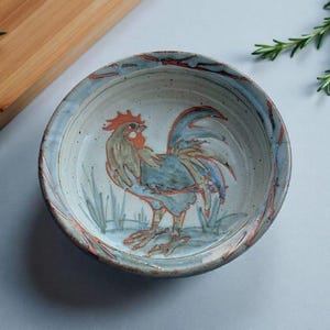 May include: A ceramic bowl with a hand-painted rooster design. The bowl has a light blue rim with red accents and a rooster in shades of blue, red, and tan. The bowl is round and sits on a light grey surface.