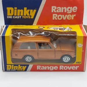 May include: A Dinky Die Cast Toys Range Rover model car in its original box. The die-cast car is a copper color with white interior details and black tires. The box is yellow, blue, and red with the Dinky and Range Rover logos.