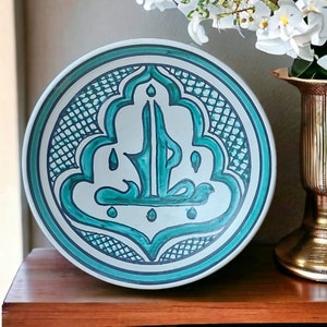 Puede incluir: Un plato decorativo de cerámica con un diseño turquesa y blanco. El plato presenta un intrincado patrón turquesa alrededor del borde y un diseño central turquesa con escritura árabe. El plato se exhibe sobre una superficie de madera.