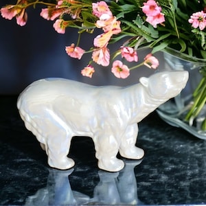 Op de afbeelding: Een parelmoerwitte ijsbeerfiguur. De beer staat op vier poten met zijn kop omhoog. Het beeldje heeft een glanzende afwerking en reflecteert licht met hints van blauw en paars. Op de achtergrond is een vaas met roze bloemen te zien.