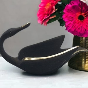 Può includere: Una ciotola decorativa a forma di cigno nero con finiture dorate. La ciotola è realizzata in un materiale nero opaco e ha un design elegante e moderno. La testa del cigno è elegantemente curva e l'interno della ciotola è di un colore scuro a contrasto. È un oggetto decorativo.