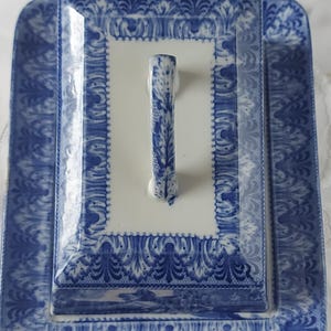 May include: A rectangular, white and blue ceramic serving dish with a lid. The dish and lid feature a detailed blue floral pattern. The lid has a vertical handle with a matching blue design. The dish is ideal for serving food.