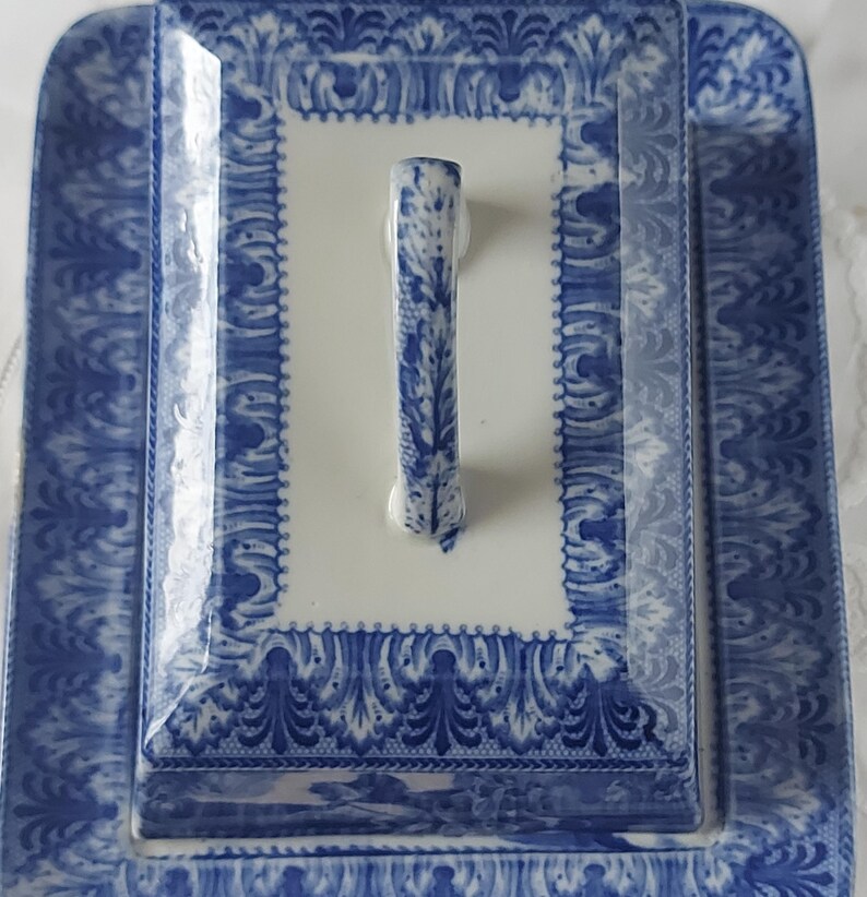 May include: A rectangular, white and blue ceramic serving dish with a lid. The dish and lid feature a detailed blue floral pattern. The lid has a vertical handle with a matching blue design. The dish is ideal for serving food.