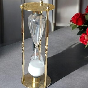 Op de afbeelding: Een decoratieve zandloper met een goudkleurig metalen frame en voet. De heldere glazen bollen bevatten wit zand. De bovenkant van de zandloper heeft een cirkelvormige plaat met een wijzerplaat. Een klassiek tijdmeetapparaat.