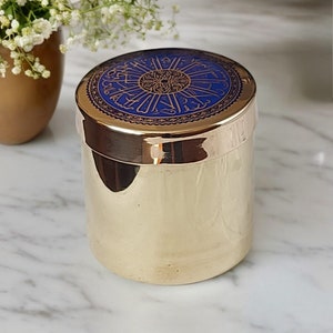Op de afbeelding: Een goudkleurige cilindrische container met een blauw deksel met goudkleurige sierlijke kalligrafie. De container heeft een glanzend, reflecterend oppervlak. Het ontwerp van het deksel is cirkelvormig, met ingewikkelde patronen. De container staat op een marmeren oppervlak.