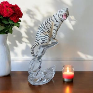 Pode incluir: Uma estatueta de tigre branco com listras pretas está em cima de uma base de cristal transparente e facetado. O tigre é retratado com a boca aberta. Uma vela vermelha em um castiçal de vidro e um vaso branco com flores vermelhas também estão presentes.
