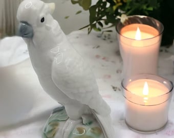 May include: A white ceramic cockatoo figurine with a gray beak and black eyes. The bird is perched on a base with green and brown accents. Two lit candles in clear glass holders are in the background, along with flowers.
