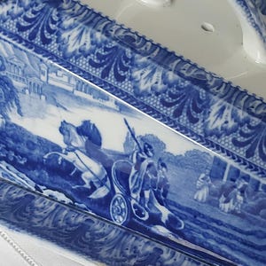 May include: A rectangular, white ceramic serving dish with a blue and white transferware design. The design depicts a classical scene with figures in a horse-drawn carriage and architectural elements. The rim features a decorative floral pattern.