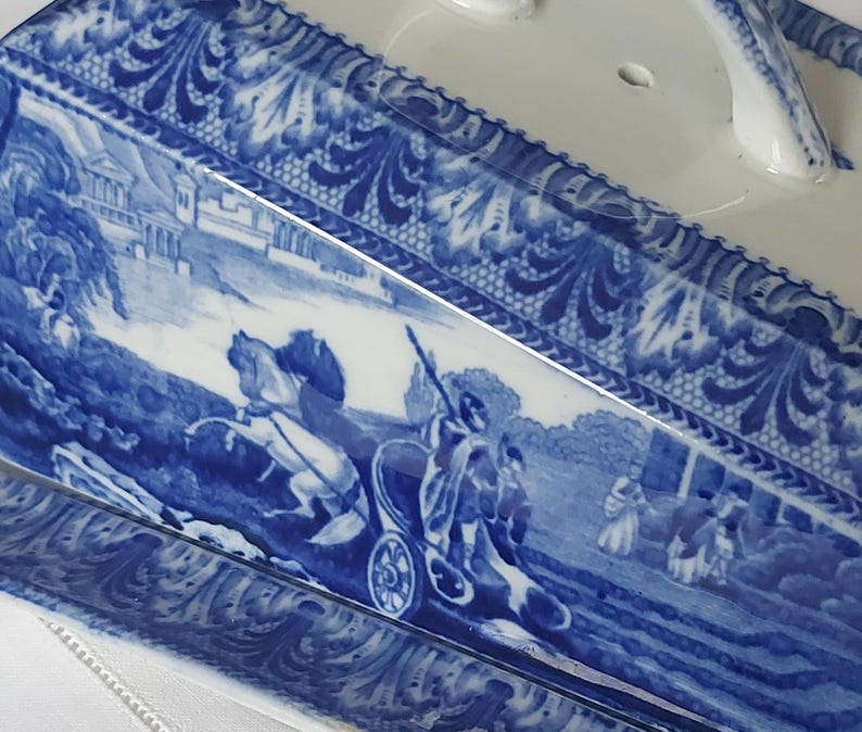 May include: A rectangular, white ceramic serving dish with a blue and white transferware design. The design depicts a classical scene with figures in a horse-drawn carriage and architectural elements. The rim features a decorative floral pattern.