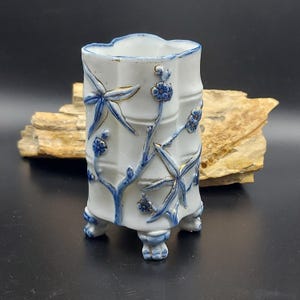 May include: A white ceramic vase with a bamboo design. The vase features blue and gold accents, with a scalloped rim and three small feet. The bamboo stalks and floral details are painted in blue.