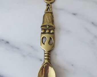 May include: A decorative brass spoon with an ornate design. The spoon features a heart-shaped top, a wheel-like center, and a bell-shaped section with intricate details. The spoon's bowl is oval-shaped and polished, reflecting light. The background includes red roses.