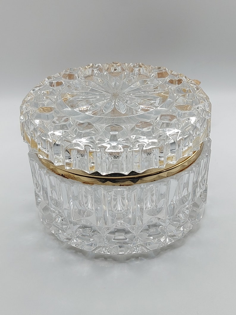 Puede incluir: Un tarro de cristal transparente y facetado con borde y tapa dorados. La tapa presenta un dise&ntilde;o central en forma de estrella. El tarro es redondo y se apoya sobre una superficie blanca. El cristal tiene un patr&oacute;n de diamante.