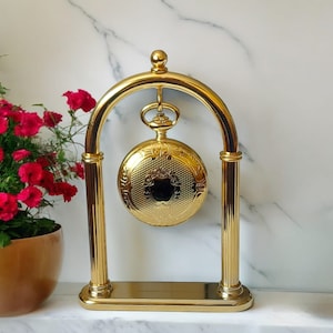 Puede incluir: Un reloj de bolsillo dorado suspendido dentro de un soporte arqueado a juego. El reloj presenta un diseño ornamentado y grabado y un centro oscuro. El soporte tiene una base rectangular y soportes verticales.