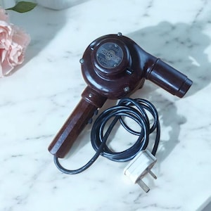 May include: A vintage brown hair dryer, branded "ORMOND", with a long handle and nozzle. A black cord and a white plug are attached. The hair dryer is positioned on a white marble surface. The design suggests a classic, retro aesthetic.