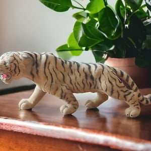 Pode incluir: Estatueta de tigre bege com listras pretas, em pose de caminhada. A boca do tigre está aberta, revelando detalhes vermelhos. A estatueta está sobre uma superfície de madeira, com plantas verdes ao fundo.