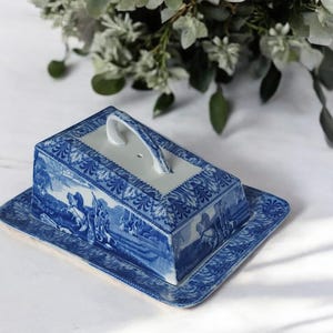 Può includere: Burriera in ceramica blu e bianca con coperchio e manico. Il piatto rettangolare e il coperchio presentano una scena paesaggistica e motivi decorativi. Un pezzo classico per la cucina, ideale per il burro.