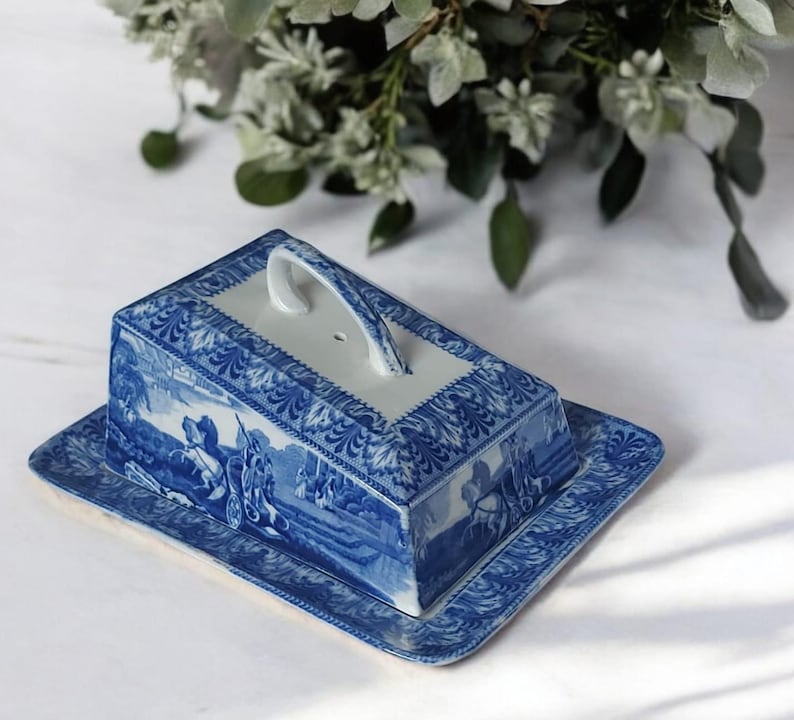 May include: Blue and white ceramic butter dish with a lid and handle. The dish features a rectangular base and a matching lid with a handle. The design includes a landscape scene and decorative patterns. A classic piece for the kitchen.