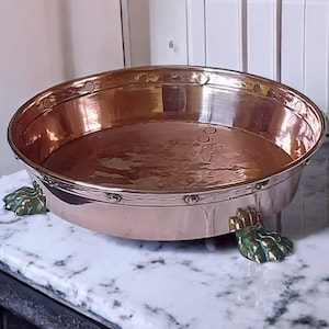 Peut inclure: Un bassin ovale en cuivre avec des rivets décoratifs sur le bord, soutenu par trois pieds ornés de couleur verte. L'intérieur reflète la lumière, suggérant un style vintage ou antique. Le diamètre du bassin est d'environ 40 cm.