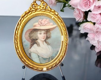 Puede incluir: Retrato ovalado antiguo en un marco dorado. El retrato representa a una mujer con un gran sombrero rosa y un vestido azul claro. El marco está sostenido por un pequeño soporte de metal. Un ramo de rosas rosas está en el fondo.