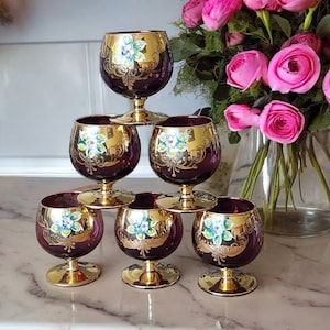Könnte beinhalten: Sechs Vintage-Cognacgläser gestapelt. Die Gläser haben eine tiefviolette Schale mit goldenen Rändern und Böden. Jedes Glas ist mit einem Blumendesign in Weiß, Blau und Grün verziert. Ein Strauß rosa Rosen ist im Hintergrund.