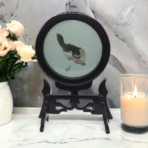May include: A circular, dark wood framed embroidery of a cat with black, white, and grey fur, set against a pale blue background. The embroidery is displayed on a black wooden stand. A lit candle in a glass holder is to the right.