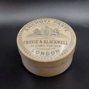 May include: A vintage Crosse & Blackwell Anchovy Paste tin. The round, off-white lid is printed with the words "ANCHOVY PASTE" and "FOR SANDWICHES & C". The text "LONDON" is also visible. The tin is on a black surface.