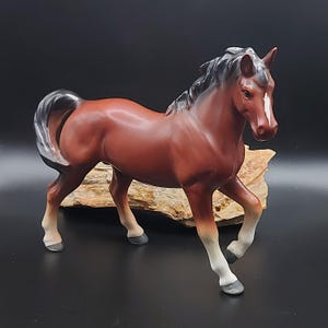 Op de afbeelding: Een keramieken paardenbeeldje in een wandelhouding. Het paard is voornamelijk bruin met een zwarte manen en staart. Het heeft witte aftekeningen op zijn gezicht en benen. Het beeldje wordt getoond op een stuk hout tegen een zwarte achtergrond.