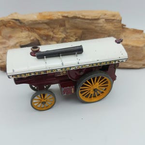 Puede incluir: Un modelo de juguete de máquina de vapor vintage, con techo blanco, carrocería granate y ruedas amarillas. Tiene una chimenea negra y el texto "LESNAY'S MOVER AMUSEMENT" es visible en el lateral. El juguete está sobre un fondo neutro.