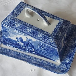 May include: Blue and white ceramic butter dish with a lid and handle. The dish features a rectangular base and a matching rectangular lid with a curved handle. The design includes a landscape scene and decorative borders.