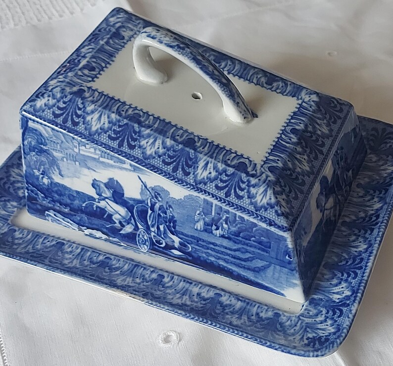 May include: Blue and white ceramic butter dish with a lid and handle. The dish features a rectangular base and a matching rectangular lid with a curved handle. The design includes a landscape scene and decorative borders.