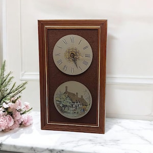 Puede incluir: Un reloj vintage con marco de madera y fondo texturizado marrón. La esfera del reloj tiene números romanos y un centro decorativo. Debajo del reloj hay una imagen circular de una casa con techo de paja. El marco es de color marrón oscuro.