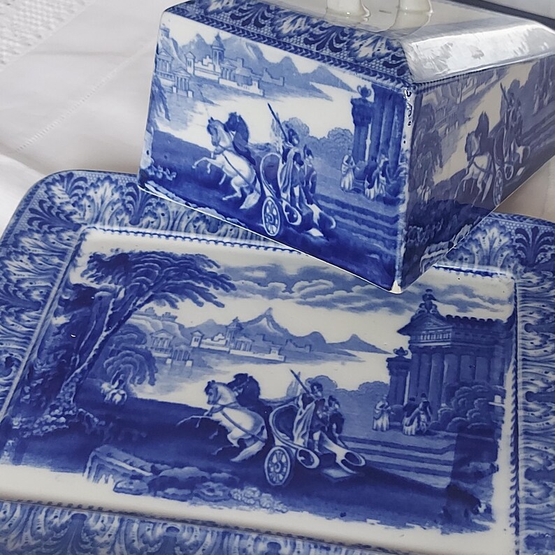 May include: Blue and white porcelain serving dish and lid with a classical scene. The rectangular dish and lid feature a detailed illustration of a chariot pulled by horses, set against a landscape with buildings and mountains. The edges have a decorative border.