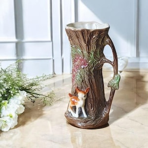 Può includere: Un vaso in ceramica marrone a forma di tronco d'albero, con manico e una piccola statuina di cerbiatto. Il vaso è decorato con elementi floreali e foglie nei toni del verde e del rosa. Il vaso è alto circa 25 cm.