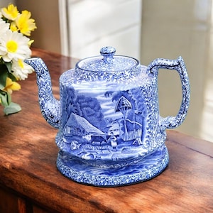 Pode incluir: Bule de cerâmica azul e branco com um design de paisagem rural. O bule apresenta uma tampa, um bico e uma alça e assenta sobre uma base a condizer. O design inclui casas, árvores e figuras. O bule é exibido numa superfície de madeira.