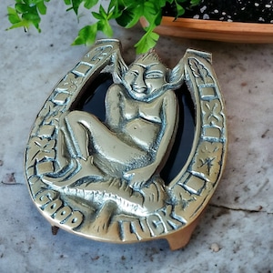 Peut inclure: Sculpture en forme de fer à cheval en laiton avec une figurine d'elfe assis. La sculpture porte l'inscription « GOOD LUCK ». L'elfe est placé sur un fond noir. Le fer à cheval est soutenu par une petite base rectangulaire.