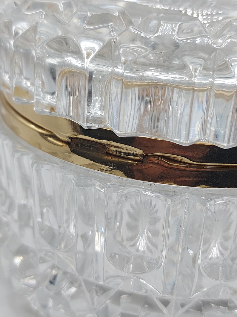 Puede incluir: Primer plano de un recipiente de vidrio transparente y facetado con una banda met&aacute;lica dorada. El vidrio tiene un patr&oacute;n de diamantes y un dise&ntilde;o floral decorativo. El recipiente es probablemente un joyero o un portador de baratijas.