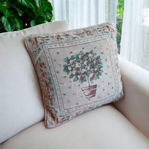 May include: Square decorative pillow with a tapestry design. The pillow features a central image of a potted tree with pink fruit, surrounded by a floral border in shades of pink, green, and beige. The pillow is placed on a white sofa.