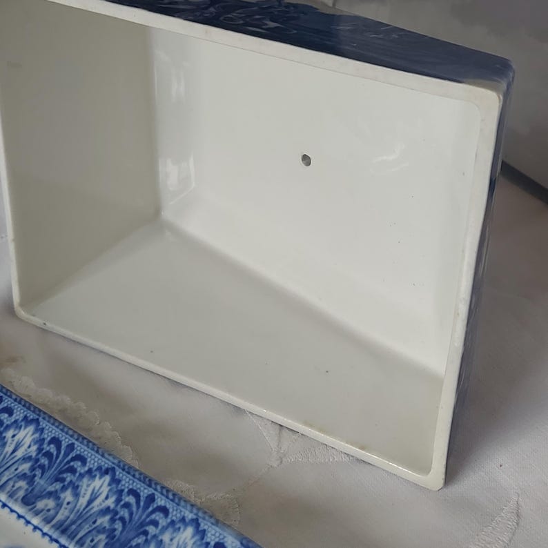 May include: A white rectangular ceramic container with a blue patterned border. The interior is plain white, and the container has a small hole on one side. The container is likely for storage or display.