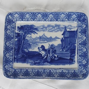 May include: Rectangular blue and white ceramic serving dish. The center features a landscape scene with a chariot, figures, and buildings. The border has a detailed, repeating floral pattern. A classic, decorative piece.