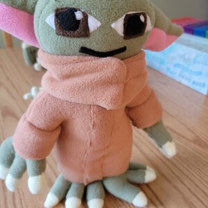 May include: A plush toy of a green alien character with large ears, brown eyes, and a pink mouth. The toy is wearing a brown hooded jacket.