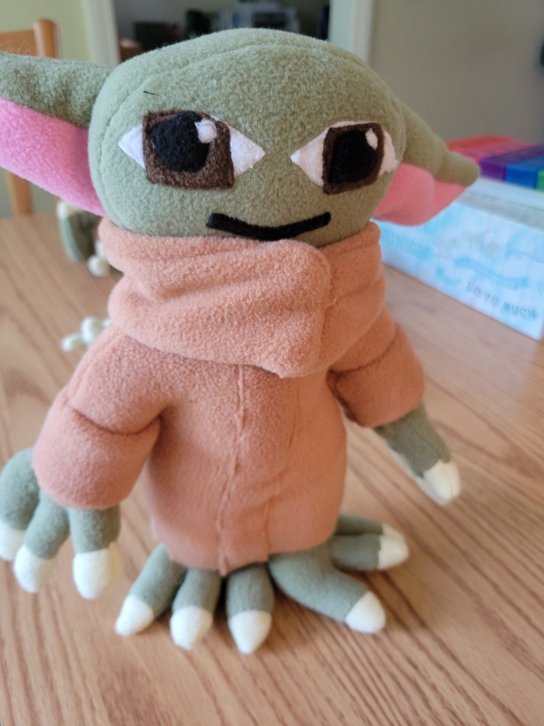 May include: A plush toy of a green alien character with large ears, brown eyes, and a pink mouth. The toy is wearing a brown hooded jacket.