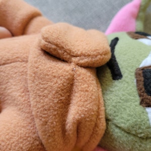 May include: A plush toy with brown, green, and pink fabric. The toy has a brown hooded head and a green body with a pink ear.